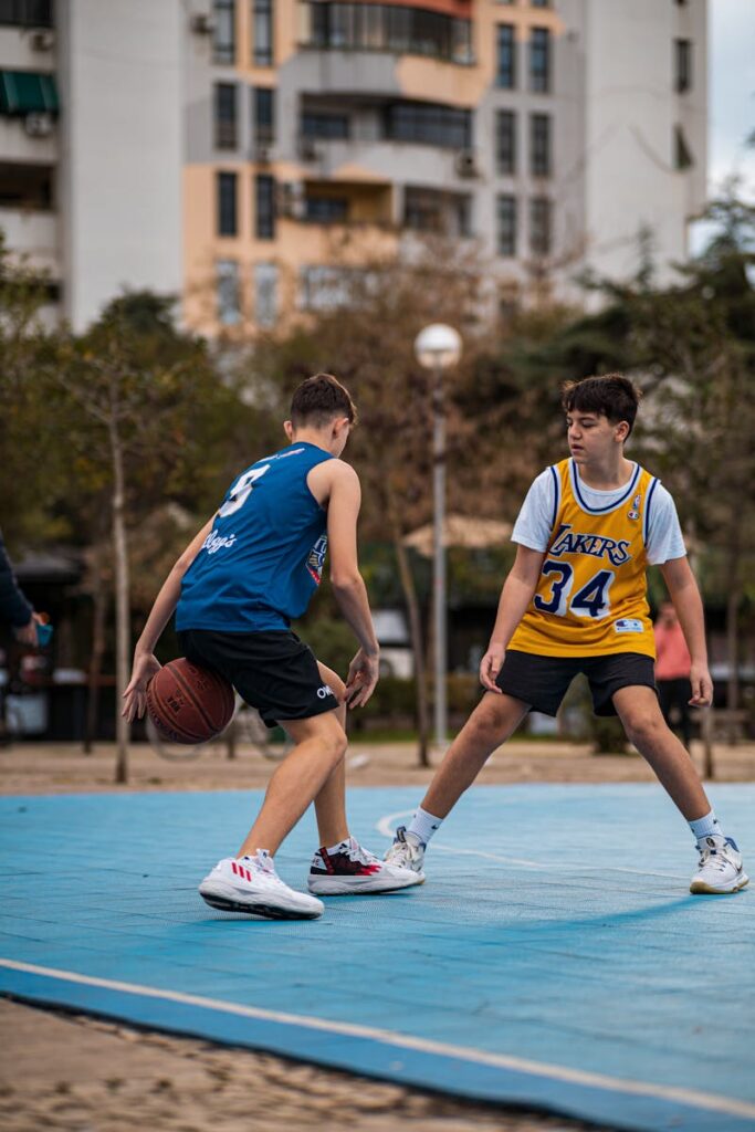 Two teenagers in sports jerseys playing basketball on an outdoor urban court.